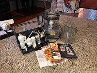Full lot showing Magimix food processor base unit, six blades and accessories arranged on a black tray, and several recipe manuals/booklets.