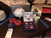 Photo showing one signed white Washington Capitals mini helmet, a display case with authentic game-used net pieces, a 2005-2006 NHL Rookie card boxed set, and three hockey pucks stacked with Capitals logo visible.