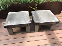 Two square side tables with cement tops and wooden legs placed on wooden deck floor with some plants in background
