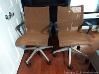Front view of two burnt orange mesh office chairs side by side showing seating area, armrests and wheeled swivel bases.