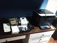 Samsung CLX 3185 printer, Canon Selphy DS810 printer, Canon SD 600 camera, Yashica T4 camera, accessories, cables, manuals and photo paper laid out on wooden surface.