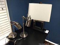 Three table lamps on black tabletop against blue wall: two brushed chrome adjustable lamps, one metal lamp with black base and white rectangular fabric shade.