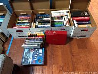 Three large cardboard boxes filled with numerous paperback and hardcover books arranged on a wood floor including some books outside the boxes such as a red large book and a hockey encyclopedia.