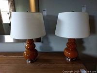 Photo showing both burnt orange glass lamps with white fabric shades and metallic bases placed side by side on wooden surface.