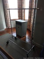 Photo showing metal rolling wardrobe rack with chrome finish and caster wheels, placed in front of window and near wall.