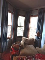 Room corner with three large windows dressed with blue fabric curtains with burnt orange trim. Beige sofa partially visible. Curtains hang from rods above windows.