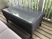 Wicker patio coffee table showing front and side angle with glass top reflecting light and wood porch floor beneath.