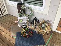 Photo showing three outdoor mats stacked on a wood deck, beige, tan, and dark blue, along with cider bottles in wooden crate, small white cabinet and decorative wreaths nearby.