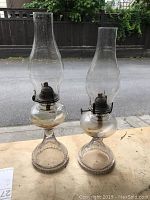 Two antique glass oil lamps standing side by side on a wooden surface outdoors, showing clear glass chimneys and bases with metal burners in between.