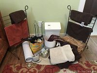 Photo showing two black metal racks with brown and cream bath towels stacked on and around them, assorted bathroom items including curling irons, toothbrush holder, Swiffer Dusters, and waste bins visible on floor.