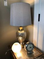 Table lamp with decorative gold-toned glass base, light gray fabric shade, small white glass globe lamp, and Westclox Big Ben alarm clock arranged on a surface.