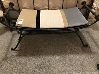 Wrought iron bench with striped cushion placed on grey carpeted floor next to a bed.