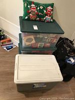Three plastic storage bins (two clear with green lids, one grey with white lid) stacked with decorative pillow on top