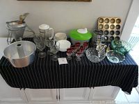 Full lot displayed on a black and white striped cloth on white cabinetry. Visible clear wine glasses, cranberry liquor glasses, white and green bowls, metal sifter, and kitchen tools including bakeware and a coffee maker.