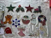 Photo showing assorted stained glass holiday window ornaments arranged on a white textured fabric, including stars, holly, teddy bear, snowman, candy cane, Christmas pudding, angel, dove, sailboat, Santa hat, and a small train ornament with a damaged hook, plus several window stickers.