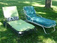Pair of vintage chaise lounges on grass, one black metal frame with green cushions and patterned bolster. Other is silver metal frame with teal button tufted cushion.