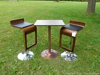 Two retro style adjustable bar stools with black cushioned wooden seats flank a small square wooden top high table on a central pedestal base, all set on grass outdoors.