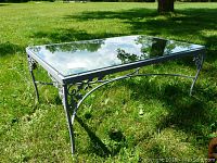 Angle showing full rectangular glass top and decorative wrought iron base with curved details and leaf motifs on an outdoor grassy surface.
