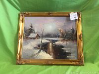 Framed oil painting showing overall winter scene with snow-covered cabin and river in ornate gold frame against green background