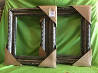 Pair of gallery wooden frames with protective corner wrapping laid side by side on a green background.