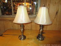 Photo of two identical brass table lamps with cream fabric shades on a wooden surface in front of a mirror.