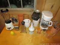 Full kitchen lot items on wooden table including blender, coffee maker, popcorn popper, knives, cheese grater, measuring cups, utensil holder, and chopper.