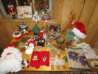 Large assortment of Christmas-themed decorations arranged on a wooden table including various figurines, several small trees, Santa hats, towels, and glass ornaments.