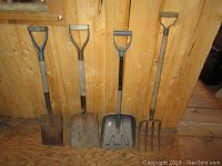 Four garden tools against wooden wall: two flat blade shovels, one black plastic scoop shovel, one metal pitch fork.
