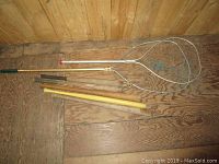Two fishing nets and baseball bats arranged on wooden floor; fishing nets with metal frames and wooden or metal handles; baseball bats mainly wooden with one plastic yellow bat