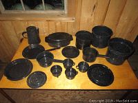 Full set of black speckled enamel cookware arranged on wooden table showing all items including pots, pans, plates, lids, and pitcher.