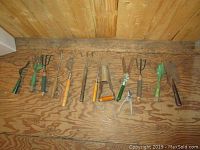 Photo showing 11 assorted gardening hand tools placed on wooden floor, arranged horizontally.