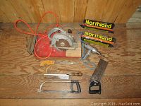 Shows power circular saw, hand saws with wooden and plastic handles, a hacksaw, electric drill, and two packs of Northland burning logs on wooden floor.