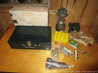 Photo showing Coleman two burner camp stove with original box, green Coleman lantern without glass, yellow plastic egg holder, Coleman electric air pump, and various smaller camping items arranged on a wooden surface.