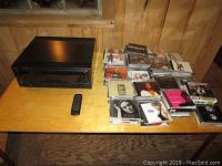 Technics CD changer and remote with a collection of 115 CDs arranged on a wooden table in a wood-paneled room.