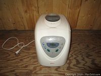 Full front view of white Sunbeam bread maker on wooden floor showing unit shape, power cord, and top viewing window.