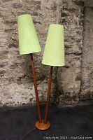 Front view of teak floor lamp with two green fabric shades against stone wall