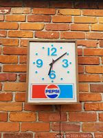 Front view of vintage Pepsi clock mounted on a brick wall showing clear blue numerals and the Pepsi logo at the bottom.