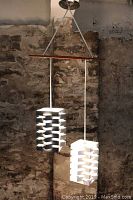 Front view of hanging light fixture with white and black geometric shades suspended from a teak bar against stone wall background