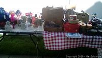 Photo showing a table with assorted vintage collectible items, tins, and beer steins arranged on a checkered red and white tablecloth and black folding tables outdoors in daylight.