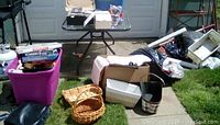 Overview of lot with purple bin showing stacks of video games, several wicker baskets on grass, various boxes, and household items.