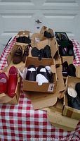 Overview of multiple pairs of women's shoes in mostly black and brown colors with one pair of bright red slip-ons, displayed in open shoe boxes on a table with a red and white checkered tablecloth.