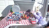 Wide view of the lot on a red and white checkered cloth showing multiple collectible items including miniature cars, a plush purple dog, a Star Wars puzzle, and a few toys.