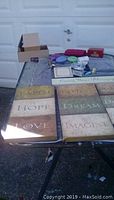 Photo showing several rectangular wall hangings with inspirational sayings 'Faith', 'Hope', 'Love', 'Believe', 'Dream', 'Imagine' plus some small boxes and statues on a table outside.