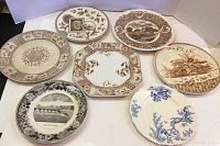 Photo showing seven antique plates from the top, displaying varied designs in blue, brown, black, and multicolor. Plates include floral, scenic, patterned, and geometric shapes.