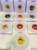 Photo showing a spread of 7-inch 45 rpm vinyl records in white paper sleeves arranged in a grid showing the record label on each disc.