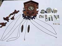 Full view of lot showing cuckoo clock body, detached carved bird pieces, two pinecone weights with chains, 3 travel alarm clocks, watch bands and parts arranged on white surface.