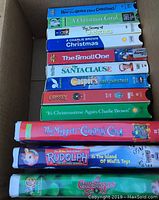 Box containing 12 colorful VHS tapes with Christmas and holiday themes, titles visible showing various classic holiday movies and shows.