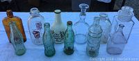 Photo showing 11 vintage glass bottles of various shapes and colors arranged on white tablecloth. Included are green Coca Cola bottles, amber bottle, Jessup Milk Bottle, clear embossed bottles, and Olive Oil bottle.
