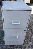 Closed view of one light gray metal file cabinet with two drawers and white handles, showing minor dents and dirt marks on top and front surfaces.