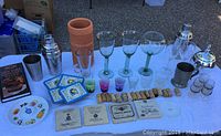 Full lot view showing assorted glassware, shot glasses, coasters, wine corks, small ice bucket, metal chalice cup, margarita salt plate, cocktail shakers, and bartender's handbook book.